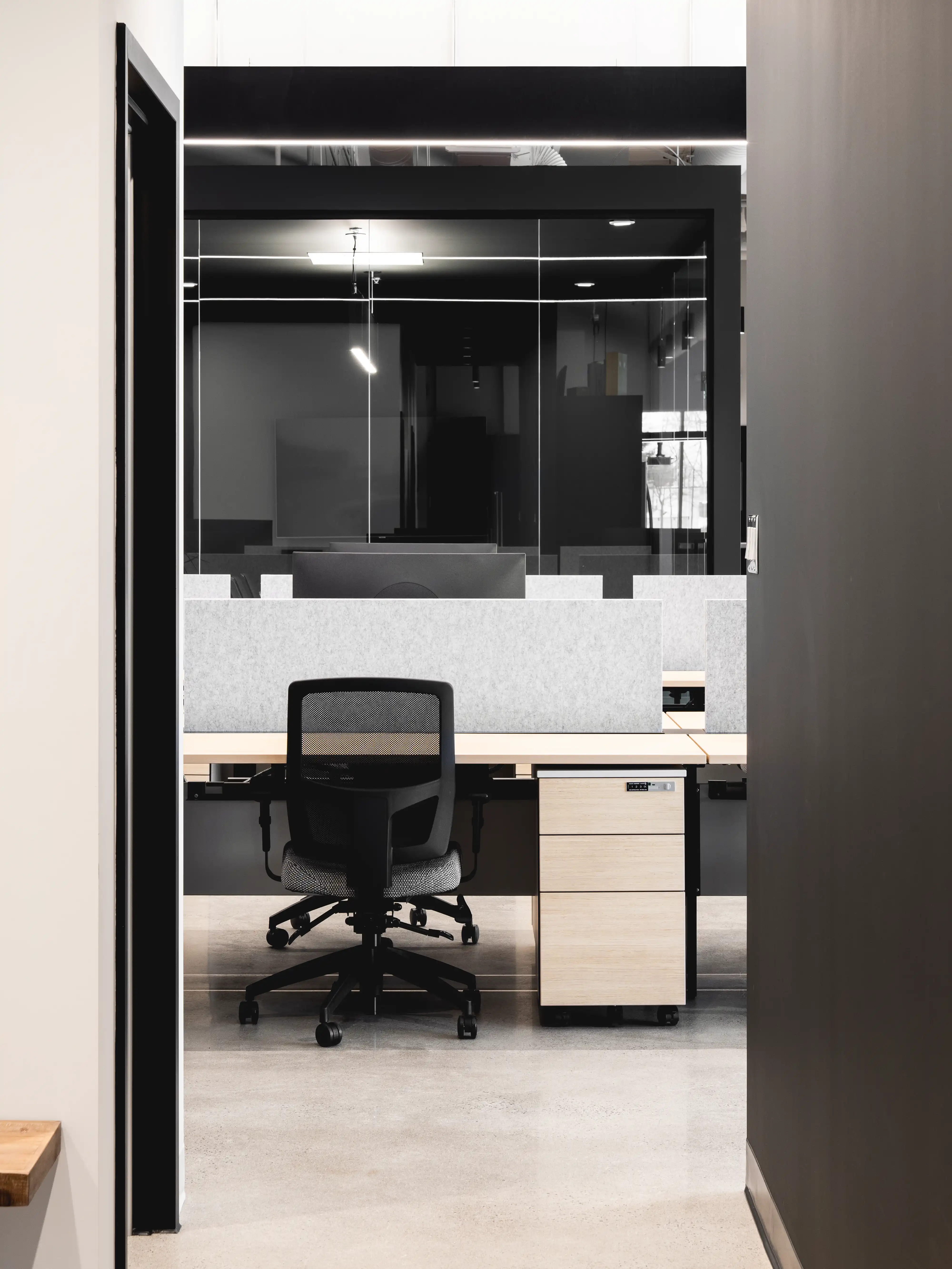 A modern office space with cubicles and a black office chair in front of a wooden desk, interior by Sarah Brown Design