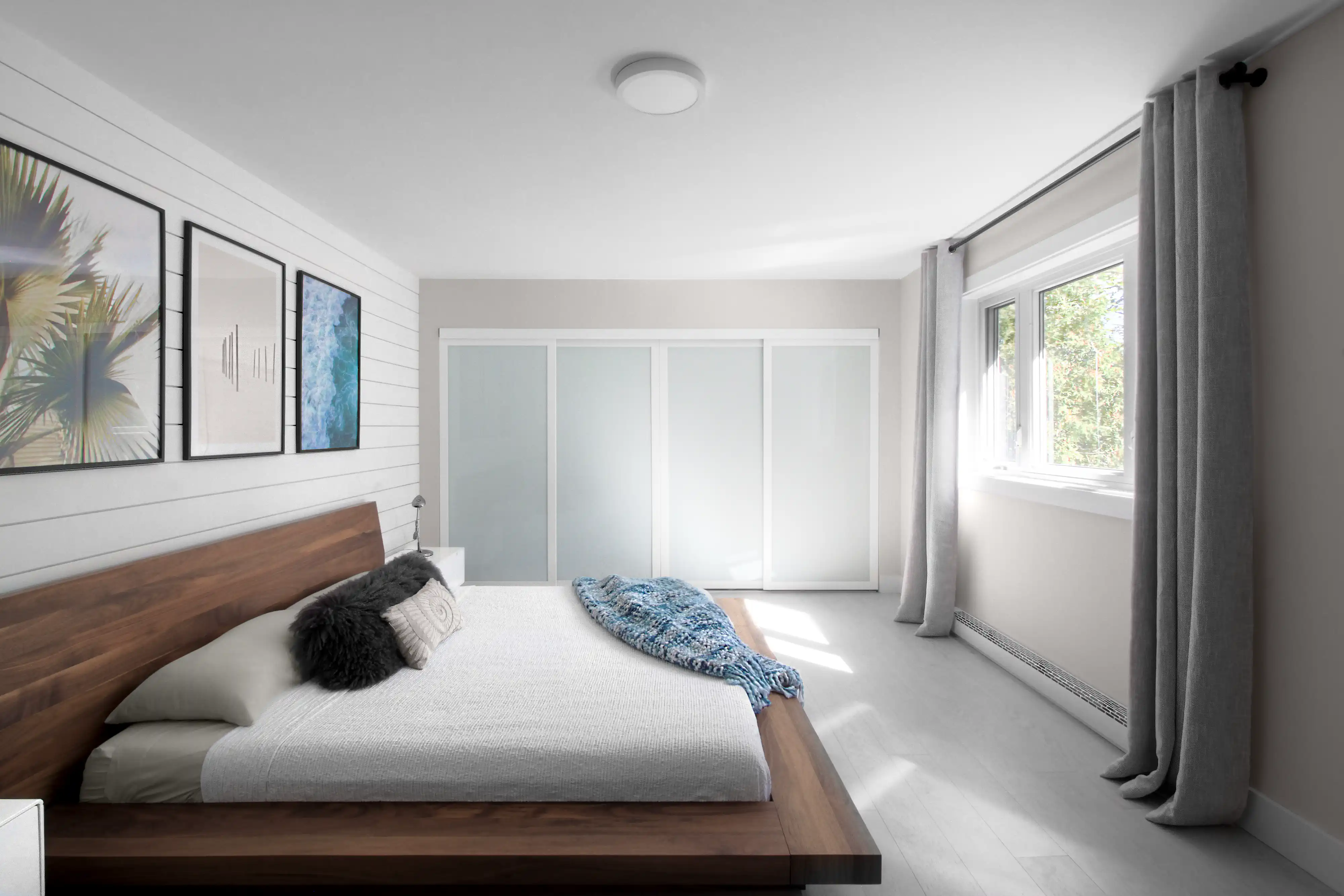 Chambre moderne avec un lit en plateforme en bois, une couverture jetée bleue, des rideaux gris et une vue sur des arbres verts, intérieur signé Sarah Brown Design