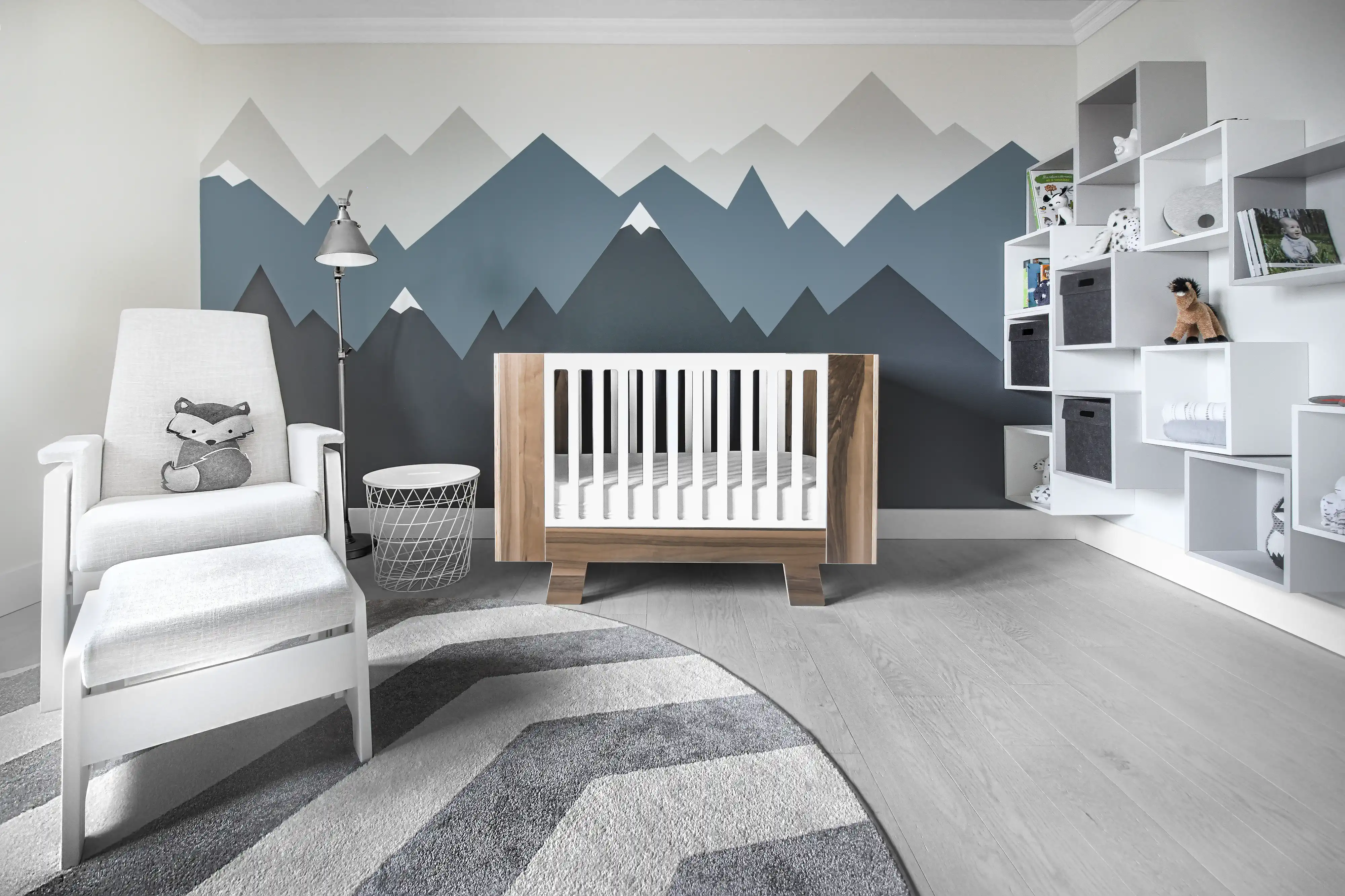 Chambre de bébé avec une fresque de montagne, un lit de bébé blanc, une chaise à bascule et une étagère remplie de jouets, intérieur signé Sarah Brown Design