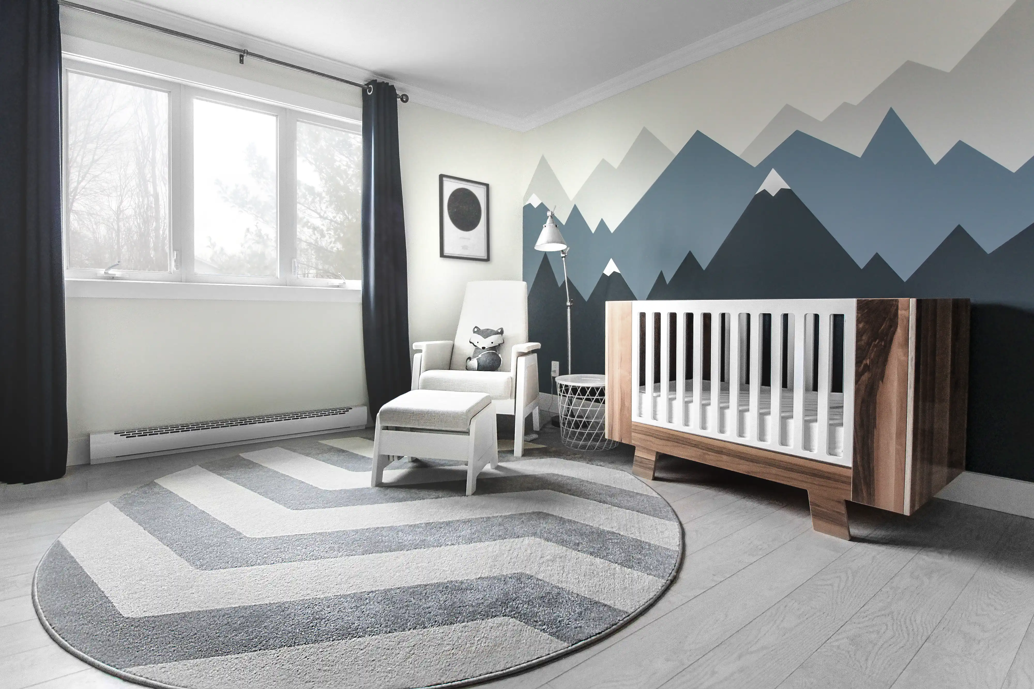 Modern nursery room with a blue and white mountain mural, a wooden crib, and a gray armchair, interior by Sarah Brown Design