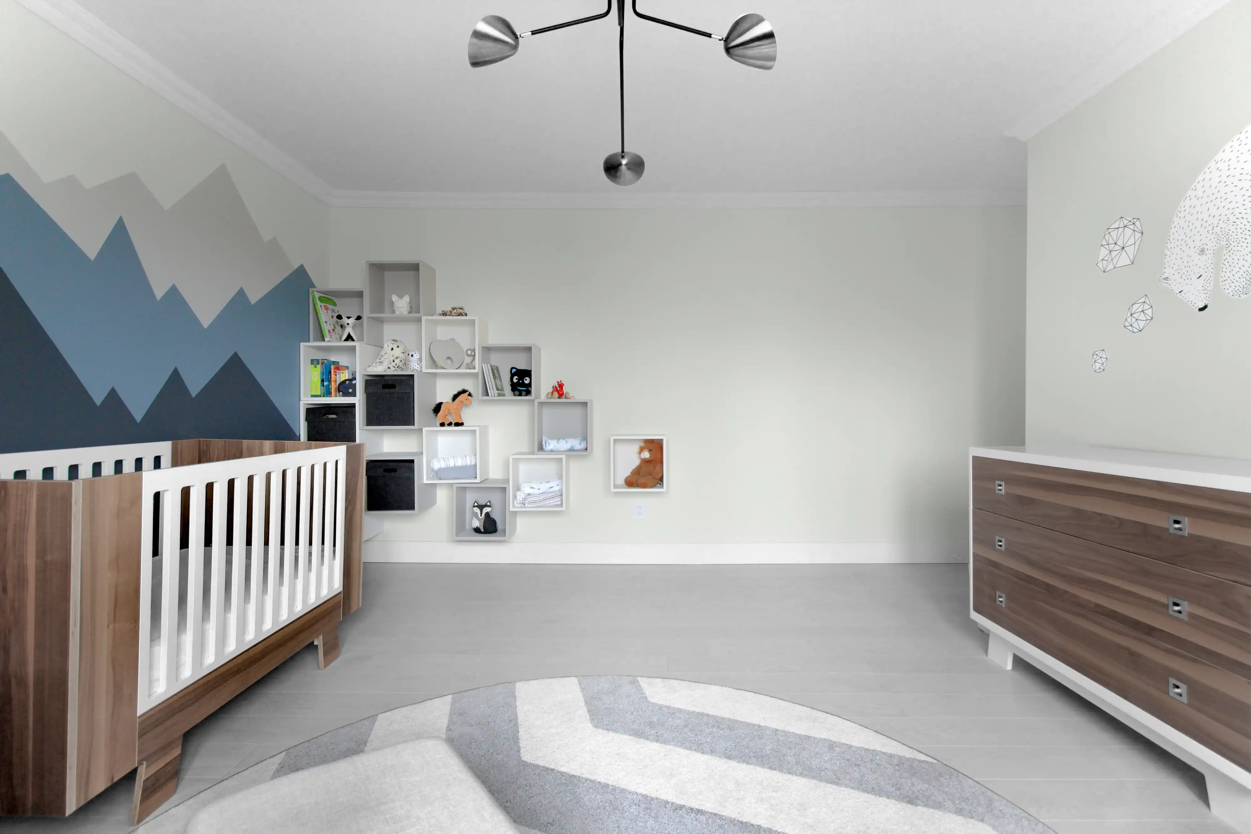 Modern nursery room with a blue and gray mountain mural, a white crib, and a wooden dresser, interior by Sarah Brown Design